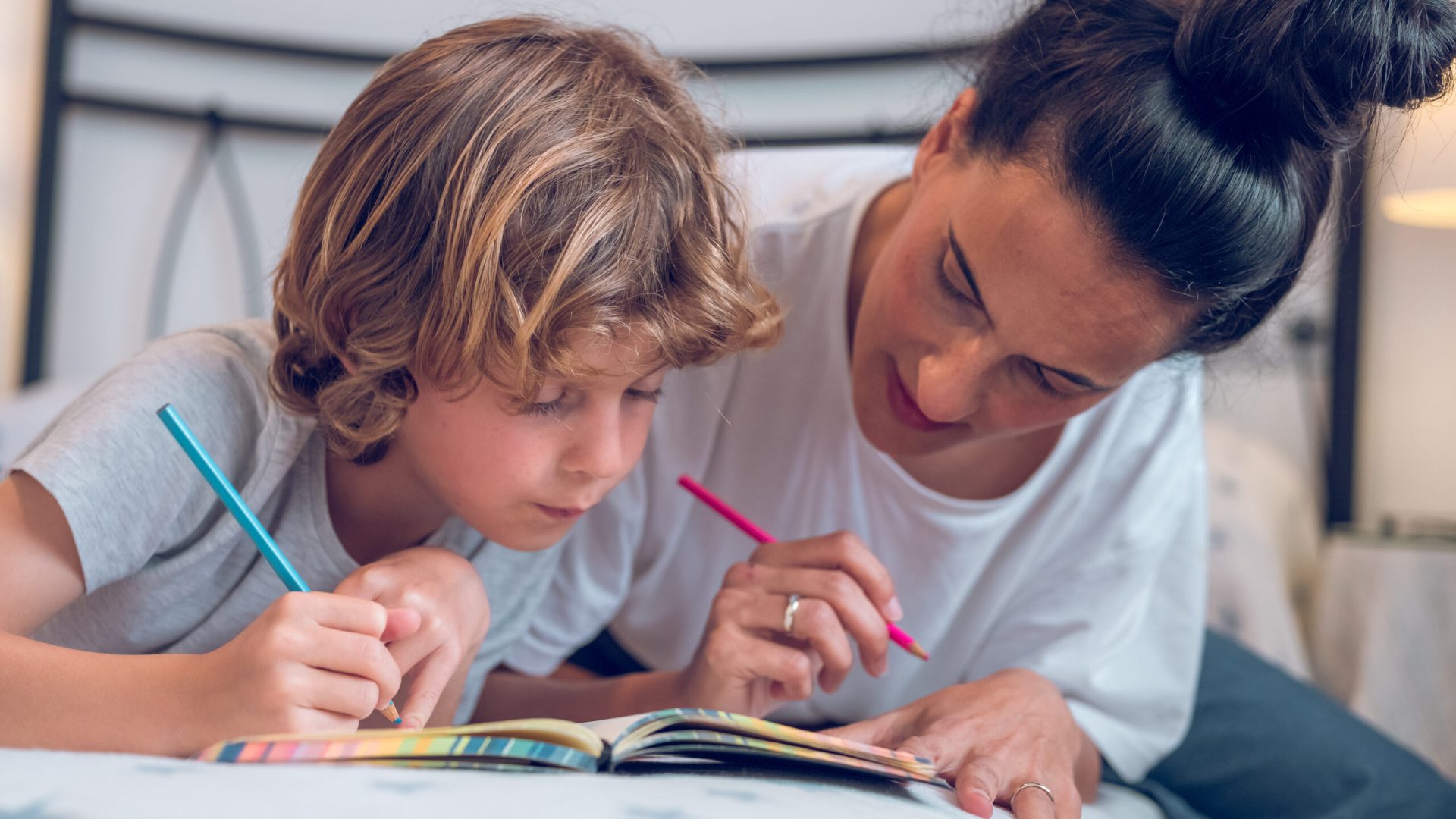 Adorable little boy painting with colorful pencils in notebook with mother while lying on bed